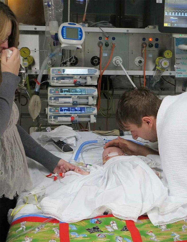 Riley, in a paediatrics intensive care unit bed connected to life support, his mother Catherine holds his hand while his father, Greg, touches his head. 