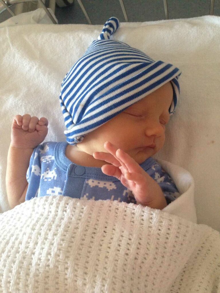 Riley, asleep under a blanket, wearing a blue and white stripped hat.