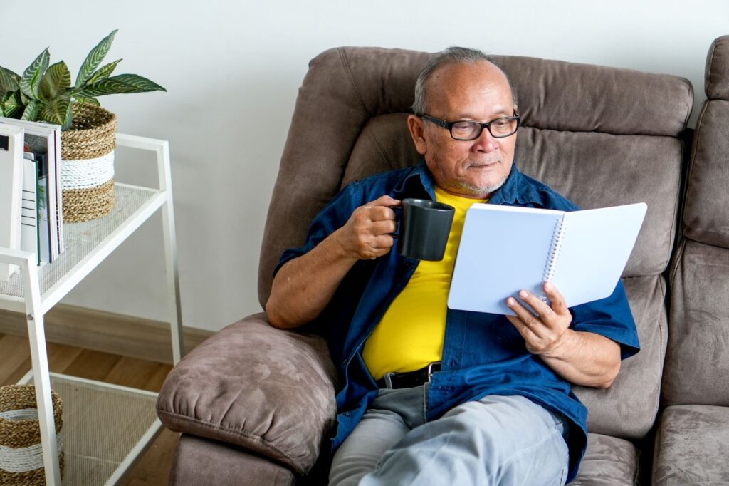 Elderly man sits on an armchair reading a book with a mug in his hand